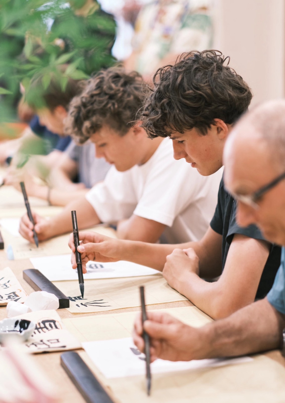 Travelers practising Chinese calligraphy with brushes at a wooden table