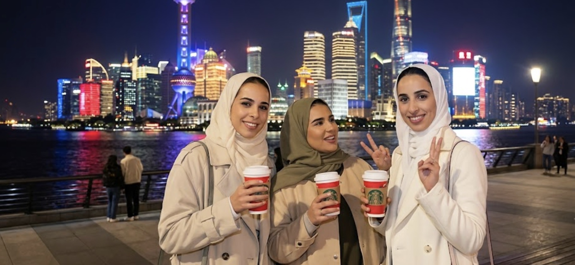 Three Muslim travellers in hijabs smiling with coffee on the Shanghai Bund at night, the lit Pudong skyline behind them