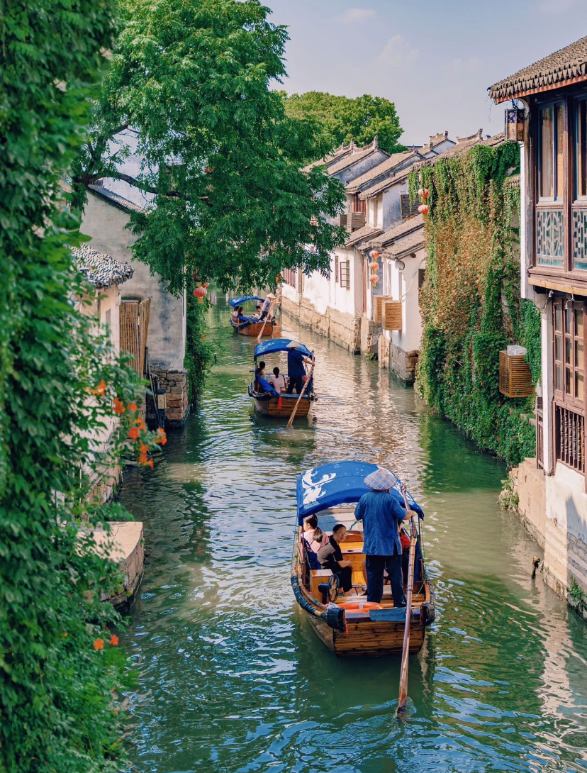 Wooden boats drifting along a tree-lined canal past whitewashed houses in a Jiangnan water town