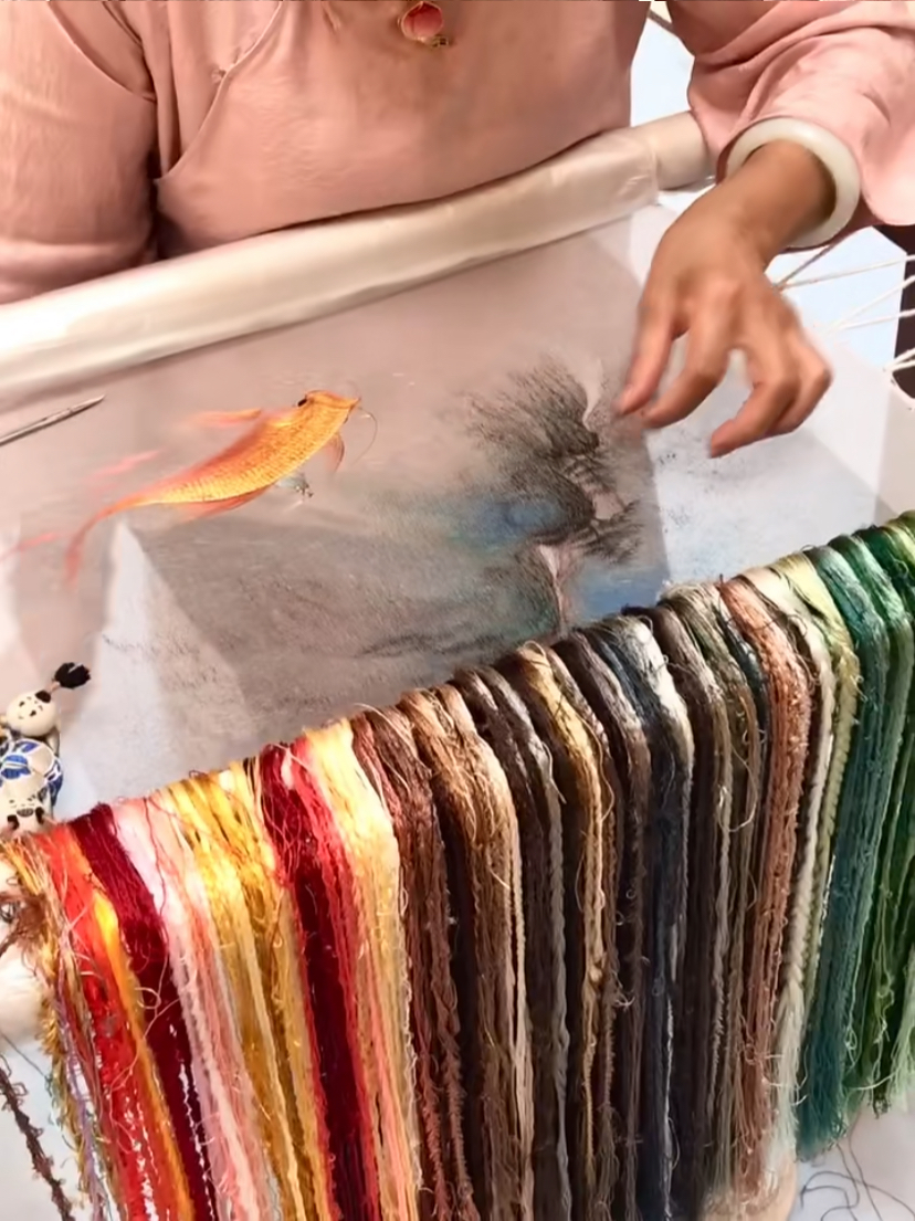 A goldfish taking shape in fine Suzhou silk embroidery beside racks of coloured thread
