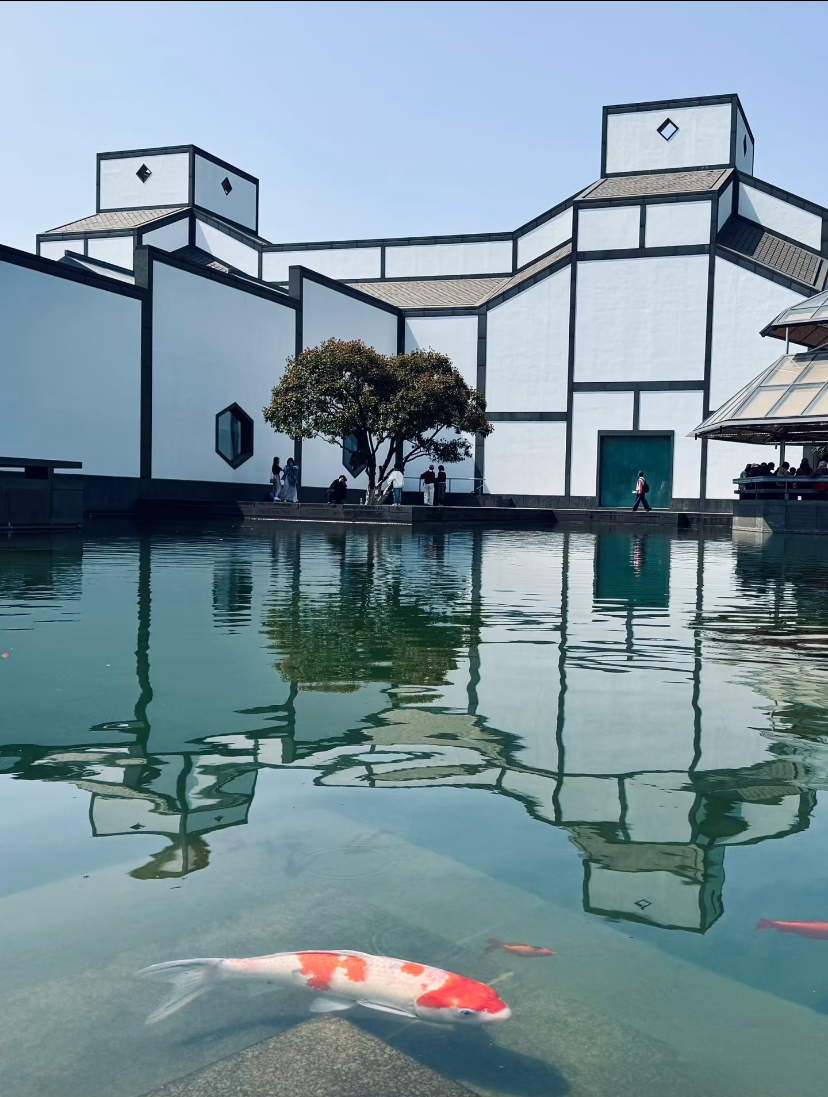 The white-and-grey geometric façade of the Suzhou Museum reflected in a koi pond